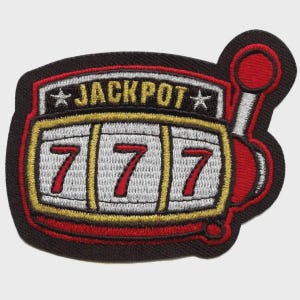 Puede incluir: Un parche bordado con forma de máquina tragaperras con borde negro. La parte superior muestra la palabra JACKPOT en amarillo, flanqueada por dos estrellas blancas. El centro presenta tres rodillos blancos con el número 7 rojo y una palanca roja.