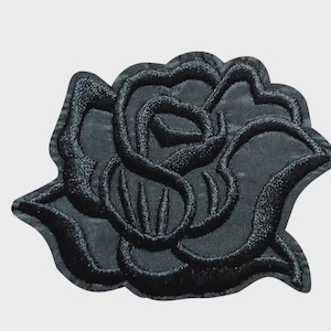 May include: A black embroidered rose patch featuring dense thread work that creates a textured, layered floral design. The edges are finished with a thick, raised black border, and the petals are defined by intricate stitching on a fabric base.