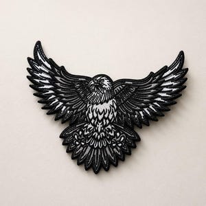 May include: An embroidered patch featuring a bald eagle in flight with wings fully spread. The design uses a monochromatic color palette of black, white, and shades of gray, with detailed stitching defining the feathers and the eagle's head.