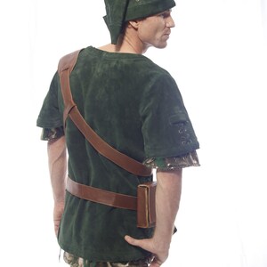 May include: A green suede tunic with brown leather straps and a matching green suede hat with a lace-up detail. The tunic has a brown leather belt and a brown leather pouch.