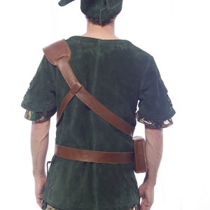 May include: A green suede tunic with brown leather straps and a brown leather belt. The tunic has a camouflage pattern on the sleeves and bottom hem. The person is wearing a green suede hat with a pointed tip.