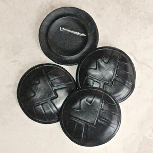 May include: Four black leather buttons with a raised design. The buttons have a pin back and are perfect for adding a unique touch to any project.