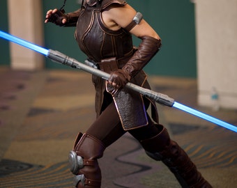 May include: A woman in a brown leather costume holds a blue lightsaber. She is wearing brown leather boots with metal accents. The photo is taken in a convention center.