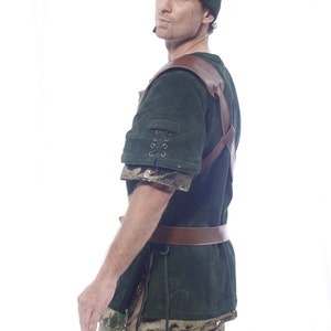 May include: A green suede tunic with brown leather straps and a matching green suede hat. The tunic has a lace-up detail on the sleeve and a brown leather belt.