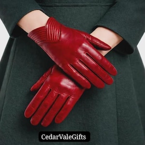 Puede incluir: Un par de guantes de cuero rojo oscuro con un acabado liso y un detalle de puño cosido en forma de V en la muñeca. Los guantes se muestran sobre un abrigo verde oscuro. El texto CedarValeGifts es visible en la parte inferior.