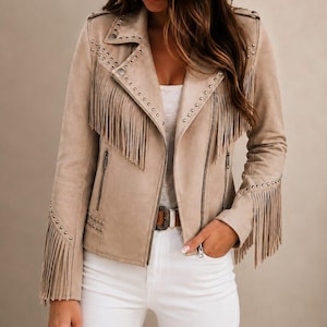 Puede incluir: Una chaqueta beige de aspecto ante con largos flecos en el pecho y las mangas, acentuada con tachuelas metálicas plateadas a lo largo de las solapas, hombros y puños. La chaqueta tiene un cierre de cremallera frontal y dos bolsillos laterales.