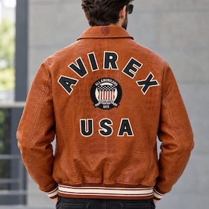 Puede incluir: Una chaqueta de cuero marrón con acabado texturizado que presenta el texto AVIREX USA en letras negras grandes en la espalda. Un parche circular en el centro muestra un gráfico de la bandera estadounidense con el texto ALL AMERICANS ESTABLISHED 1975.