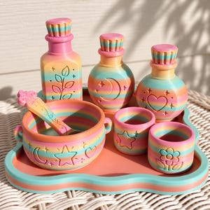 May include: A set of three rainbow-striped bottles, a cauldron with a spoon, and two small cups on a matching tray. Each item features embossed designs including a plant, moon, heart, star, and flower, all rendered in a gradient of pastel colours.