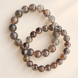 Puede incluir: Dos pulseras elásticas compuestas por cuentas redondas y pulidas. Las cuentas son translúcidas con una mezcla de inclusiones de color gris oscuro, negro y ámbar, creando una apariencia moteada en cada piedra de las piezas de joyería circulares.