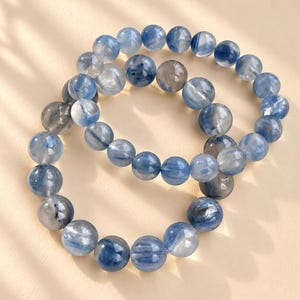 Könnte beinhalten: Zwei elastische Armbänder aus runden, polierten Kyanit-Perlen. Die Perlen haben ein durchscheinendes Aussehen mit verschiedenen Blautönen, die von hellem Himmelblau bis zu tiefem Marineblau reichen, durchsetzt mit grauen und weißen Einschlüssen.