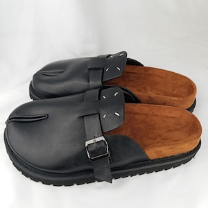 May include: A pair of black leather slip-on mules featuring a split-toe design, a wide strap across the top with a silver-toned metal buckle, and a brown suede footbed. The shoes have a thick black lug sole and four white stitches on the upper.