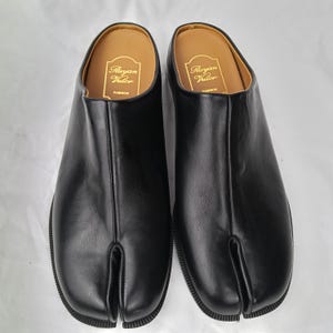 May include: A pair of black slip-on shoes with a split-toe design. The interior features a tan insole with the gold-printed text Ruyan Vutor FASHION. The shoes have a smooth black exterior finish and a textured black sole, providing a sleek look.