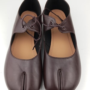 Könnte beinhalten: Ein Paar dunkelbraune Schuhe mit geteilter Zehenpartie, glatter Oberfläche, niedrigem Schnitt und Schnürung an der Oberseite. Das Innere ist mit einer beigen Innensohle ausgestattet und das Design weist eine Trennung zwischen dem großen Zeh auf.