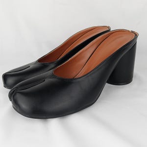 May include: A pair of black slip-on mules featuring a distinct split-toe design. These shoes have a smooth black exterior, a tan interior lining, and a sturdy block heel. The construction includes a rounded opening and a seam running down the center.
