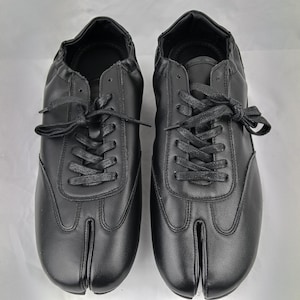May include: A pair of black lace-up shoes featuring a distinct split-toe design. The shoes have a smooth, matte finish, matching black laces, and a low-top silhouette with a rounded heel collar. The construction includes visible stitching details.