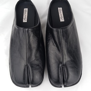 Pode incluir: Um par de sapatos sem atacadores em pele preta com um design de biqueira dividida. A palmilha interior é preta e apresenta uma etiqueta retangular branca com o texto "Happy feeling" impresso em letra cursiva preta. O couro é texturizado.