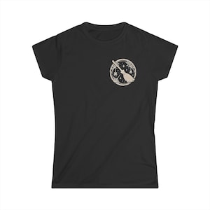 May include: A black short-sleeve t-shirt featuring a circular graphic on the left chest. The design depicts a beige broomstick centered within a frame of spiderwebs, small spiders, and stars, rendered in a light, neutral line art style.