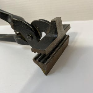 May include: A pair of black metal pliers featuring a wide, flat, T-shaped head with a textured, grooved gripping surface. The tool includes a central pivot point and a small coiled spring mechanism located between the handles for tension control.