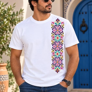 May include: A white short-sleeved t-shirt featuring a vertical, colourful cross-stitch pattern on the left side. The design consists of repeating floral and geometric motifs in shades of purple, pink, teal, and yellow, arranged in a column.