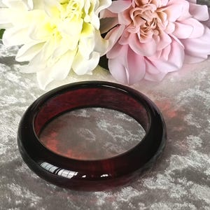Pode incluir: Uma pulseira circular lisa feita de um material vermelho escuro translúcido com um marmoreado interno subtil e uma superfície polida e reflexiva que capta a luz ao longo da sua borda exterior arredondada.