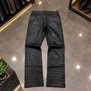 May include: Black trousers featuring a textured crocodile skin pattern throughout the fabric. The trousers include a standard waistband with a button closure, front pockets, and a straight-leg cut, presenting a uniform dark finish across the garment.