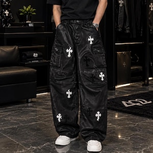Könnte beinhalten: Schwarze Cargohose mit weitem Bein und lederartiger Oberfläche. Die Hose ist mit mehreren weißen kreuzförmigen Aufnähern und dezenten, tonalen Kreuzmustern verziert. Sie verfügt über seitliche Cargotaschen und einen geknöpften Hosenschlitz.