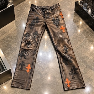 Könnte beinhalten: Dunkelbraune Lederhose mit Knopfleiste und zahlreichen dekorativen Aufnähern in Schwarz und leuchtendem Orange. Die Applikationen in Form von Kreuzen und Lilien sind auf die Vorderseite der Beine und die Taschen aufgenäht.