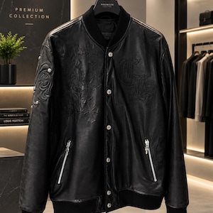 May include: A black leather varsity-style jacket featuring a ribbed collar, cuffs, and hem. The front displays embossed cross patterns and gothic-style lettering, complemented by silver-toned snap buttons and two zippered pockets with metallic pulls.