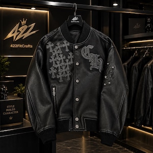 May include: A black leather varsity jacket featuring a ribbed collar, cuffs, and hem. The front displays a cluster of embroidered grey crosses on the right side, a large gothic-style monogram on the left, and silver-toned metal snap buttons.