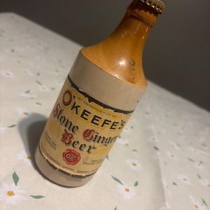 May include: A vintage O'Keefe's Stone Ginger Beer bottle with a tan ceramic body and a brown glazed neck. The label features the text O'Keefe's Stone Ginger Beer, Net Contents 10 Fluid Ozs, and O'Keefe's Beverages Limited, Toronto, Canada.