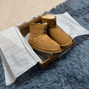 May include: A pair of tan suede ankle boots with cream-colored shearling lining, featuring a silver-toned cross-shaped metal charm attached to the front of each boot. The boots are presented inside an open cardboard box with branded tissue paper.