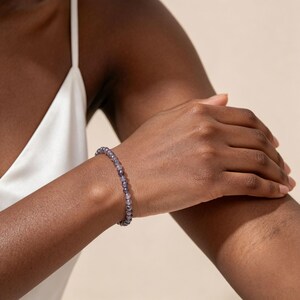 May include: A beaded bracelet featuring small, round, translucent purple stones strung together in a continuous loop. The beads have a smooth, polished finish and a consistent size, creating a simple and uniform circular band around the wrist.
