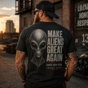May include: A black short-sleeved t-shirt featuring a detailed grey alien head graphic on the back. The text reads "MAKE ALIENS GREAT AGAIN" in large letters, followed by "EARTH TOUR 2026" and the smaller phrase "I WANT TO BELIEVE... WE CAN WIN."