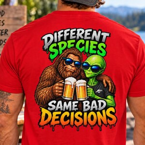 May include: A bright red t-shirt featuring a graphic on the back of a brown Bigfoot and a green alien wearing sunglasses, each holding a glass of beer. The text reads DIFFERENT SPECIES SAME BAD DECISIONS in bold, colorful, dripping-style lettering.