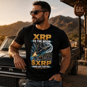 May include: A black short-sleeved t-shirt featuring a graphic print with the text XRP TO THE MOON and $XRP BANKS CAN'T STOP THIS. The design depicts a gorilla riding a rocket toward a moon, with city buildings and a bear in the foreground.