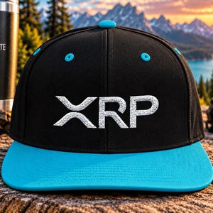 May include: A black baseball cap with a bright blue flat brim and a matching blue top button. The front of the cap features the white embroidered text XRP. The cap has blue eyelets on the crown and a structured six-panel design with stitching.