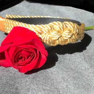 May include: A gold rope headband with a red rose lying on a grey surface.