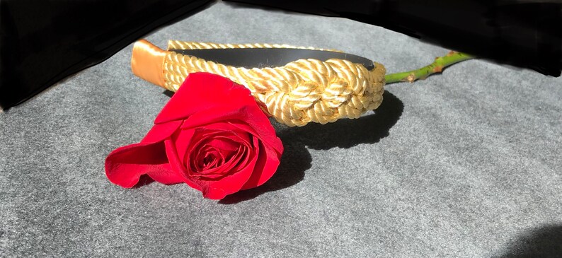 May include: A gold rope headband with a red rose lying on a grey surface.