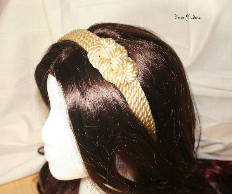 May include: A gold rope headband with a knotted design. The headband is worn on a model with long, dark brown hair.