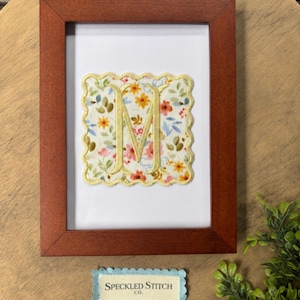 Könnte beinhalten: Ein rechteckiger brauner Holzrahmen mit weißem Hintergrund und einem mittig platzierten quadratischen Stoffaufnäher, der einen gelben Wellenrand und ein Blumenmuster mit einem großen goldenen Buchstaben M aufweist. Unter dem Rahmen befindet sich ein kleines blaues gewelltes Etikett mit dem Text Speckled Stitch co.