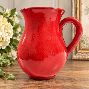 May include: A vibrant red ceramic pitcher with a glossy, smooth finish. The vessel features a wide, flared rim, a rounded body, and a sturdy, curved handle on the right side. The surface displays subtle variations in the deep red glaze throughout.