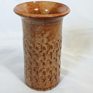 May include: A cylindrical ceramic vase with a flared rim, featuring a textured exterior surface with vertical indentations and a mottled brown and tan glaze. The vessel has a smooth, rounded top edge and a straight body that tapers slightly toward the base.