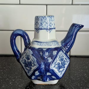 May include: A small ceramic teapot featuring a white base with intricate blue floral patterns and geometric borders. The vessel has a curved handle, a slender spout, and a cylindrical neck, all decorated with detailed blue and white motifs.