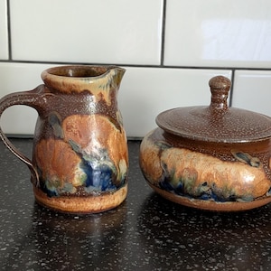 May include: A ceramic creamer pitcher and a matching lidded sugar bowl featuring a mottled glaze in shades of brown, tan, and deep blue. The creamer has a curved handle and a pouring spout, while the sugar bowl has a round lid with a knob.