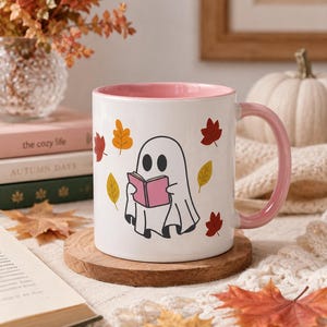 May include: A white ceramic mug with a pink interior and handle, featuring a graphic of a white ghost reading a pink book surrounded by scattered red, orange, and yellow autumn leaves. The mug has a smooth finish and a cylindrical shape.