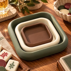 May include: A set of three nested square trays with rounded corners, featuring a dark olive green outer tray, a cream-colored middle tray, and a dark brown inner tray, all stacked together to create a compact, layered, and functional storage unit.