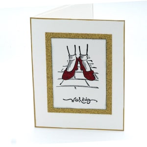 May include: A white card with a gold border and a gold glitter frame. Inside the frame is a black and white illustration of red shoes on a yellow brick road. The text "Wish Big" is written in black cursive below the illustration.