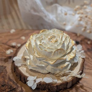 May include: A sculpted white flower with gold-painted edges on its petals, resting on a circular wooden slice. The flower features layered, textured petals with metallic gold accents on the tips, creating a detailed floral appearance.