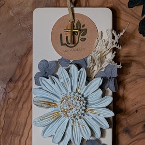 Puede incluir: Una tableta de cera rectangular de color blanco roto con una gran margarita azul claro y detalles en pan de oro. Está decorada con pétalos de hortensia azules secos, ramitas blancas, un dije de cruz dorada y una etiqueta circular LumiLeaf.Lab.