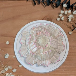 May include: A circular white tray holding a sculpted flower ornament. The flower features layered, ruffled petals in a soft beige tone with delicate pink edges and a textured, detailed center, creating a dimensional floral appearance.
